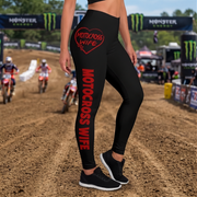 Black high-waisted leggings with red 'MOTOCROSS WIFE' text and a red heart graphic on the hip, shown on a model on a dirt motocross track with Monster Energy banners in the background.