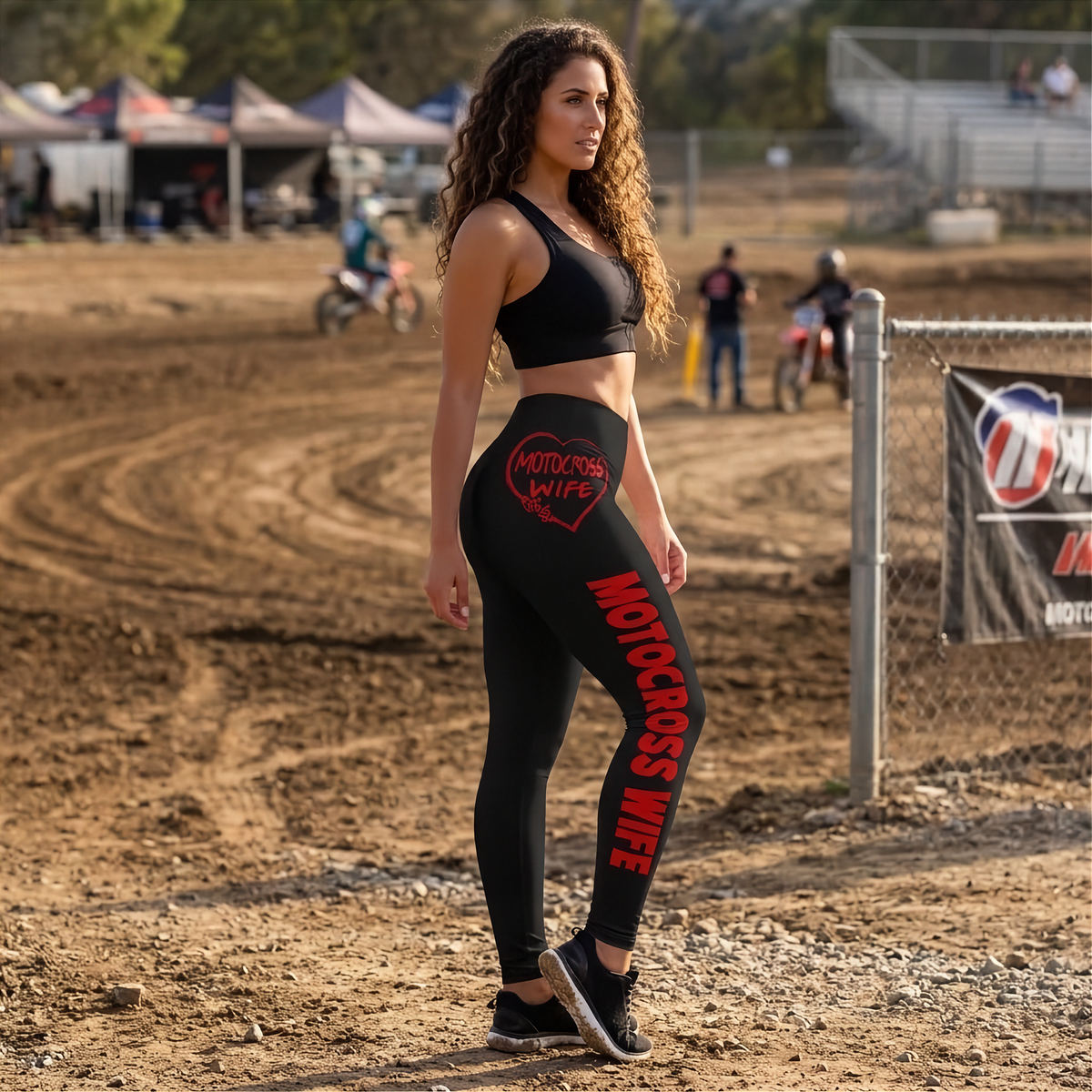 A woman stands on a dirt motocross track wearing black athletic leggings with bold red 'MOTORCROSS WIFE' text and a red heart logo on the thigh, paired with a black sports bra.