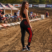 A woman stands on a dirt motocross track wearing a black sports bra and matching black leggings with bold red 'Moto Cross Wife' text and a heart graphic on the hip. Dirt bikes, tents, and track signage are visible in the background.