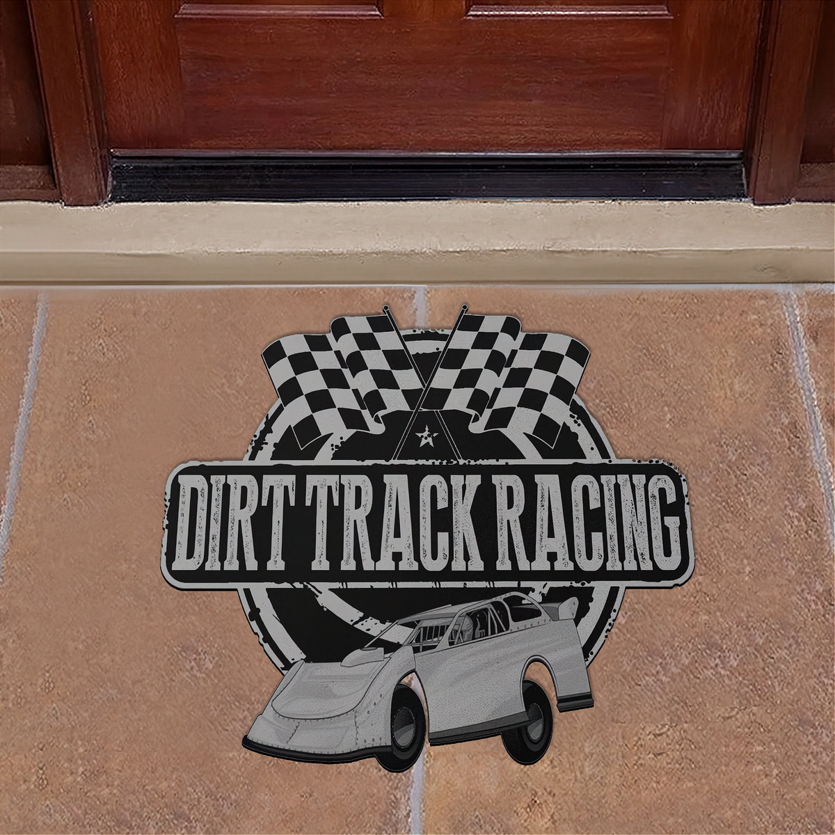 A rectangular metal doormat featuring a 'Dirt Track Racing' design, placed on a tan stone tile floor in front of a dark brown wooden door. The design includes a black and white checkered flag motif, a white race car, and distressed black lettering.