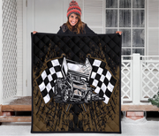 A person stands on a snowy porch holding up a large quilted blanket or throw. The blanket features a sprint car racing design with two black-and-white checkered flags, a silver hot rod car, and a distressed brown and black background.