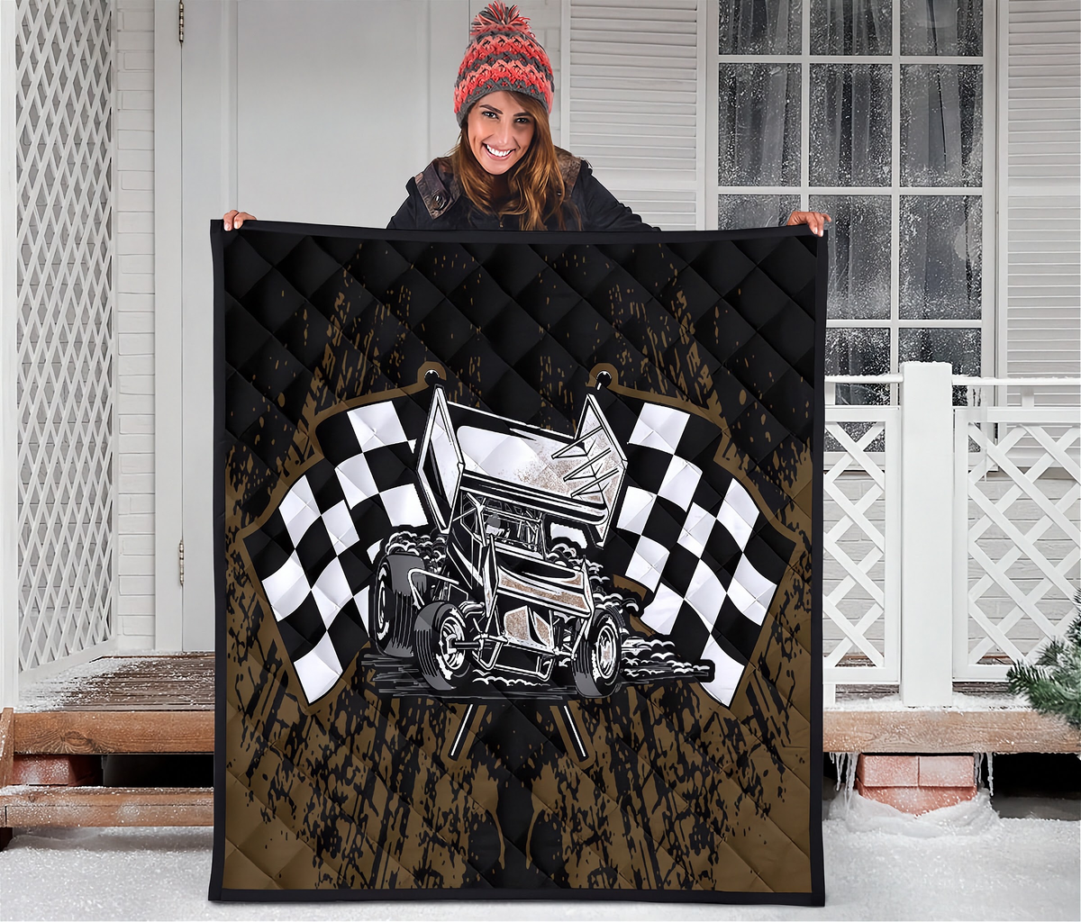 A person stands on a snowy porch holding up a large quilted blanket or throw. The blanket features a sprint car racing design with two black-and-white checkered flags, a silver hot rod car, and a distressed brown and black background.