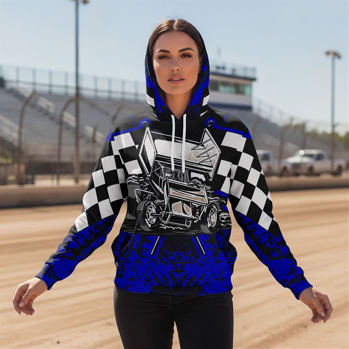 A woman stands on a dirt racetrack wearing a hooded sweatshirt with a bold racing graphic. The hoodie features a black and white checkered pattern on the sleeves, a detailed illustration of a race car on the chest, and bright royal blue sections at the hood, cuffs, and lower body.