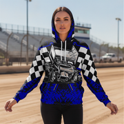 A woman stands on a dirt racetrack wearing a hooded sweatshirt with a bold racing graphic. The hoodie features a black and white checkered pattern on the sleeves, a detailed illustration of a race car on the chest, and bright royal blue sections at the hood, cuffs, and lower body.