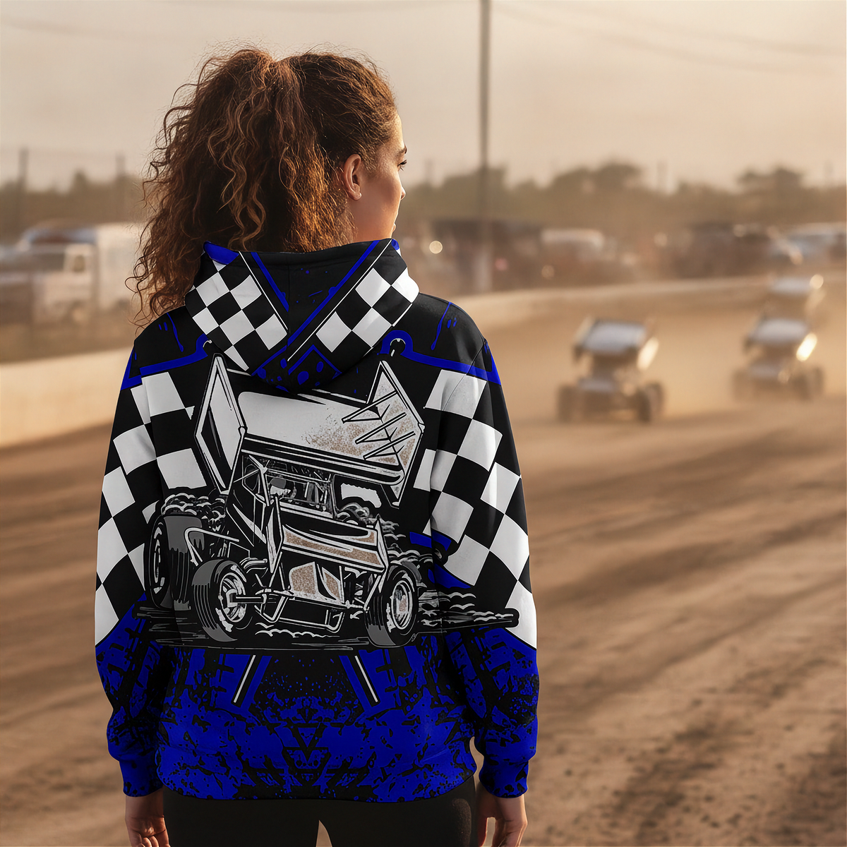 A woman stands on a dirt racetrack wearing a hooded sweatshirt with a bold racing graphic. The hoodie features a black-and-white checkered pattern on the shoulders and upper sleeves, a large illustrated sprint car on the back, and vivid royal blue lower sections with a dark abstract pattern.