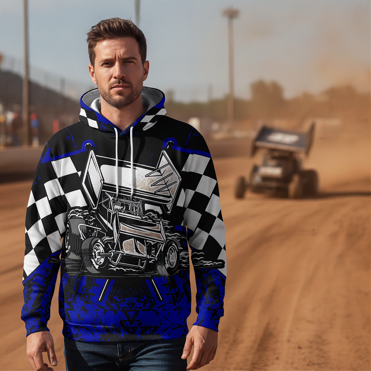 A man wearing a blue, black, and white graphic hoodie with a race car and checkered flag design, standing on a dirt racetrack with a blurred sprint car in the background.