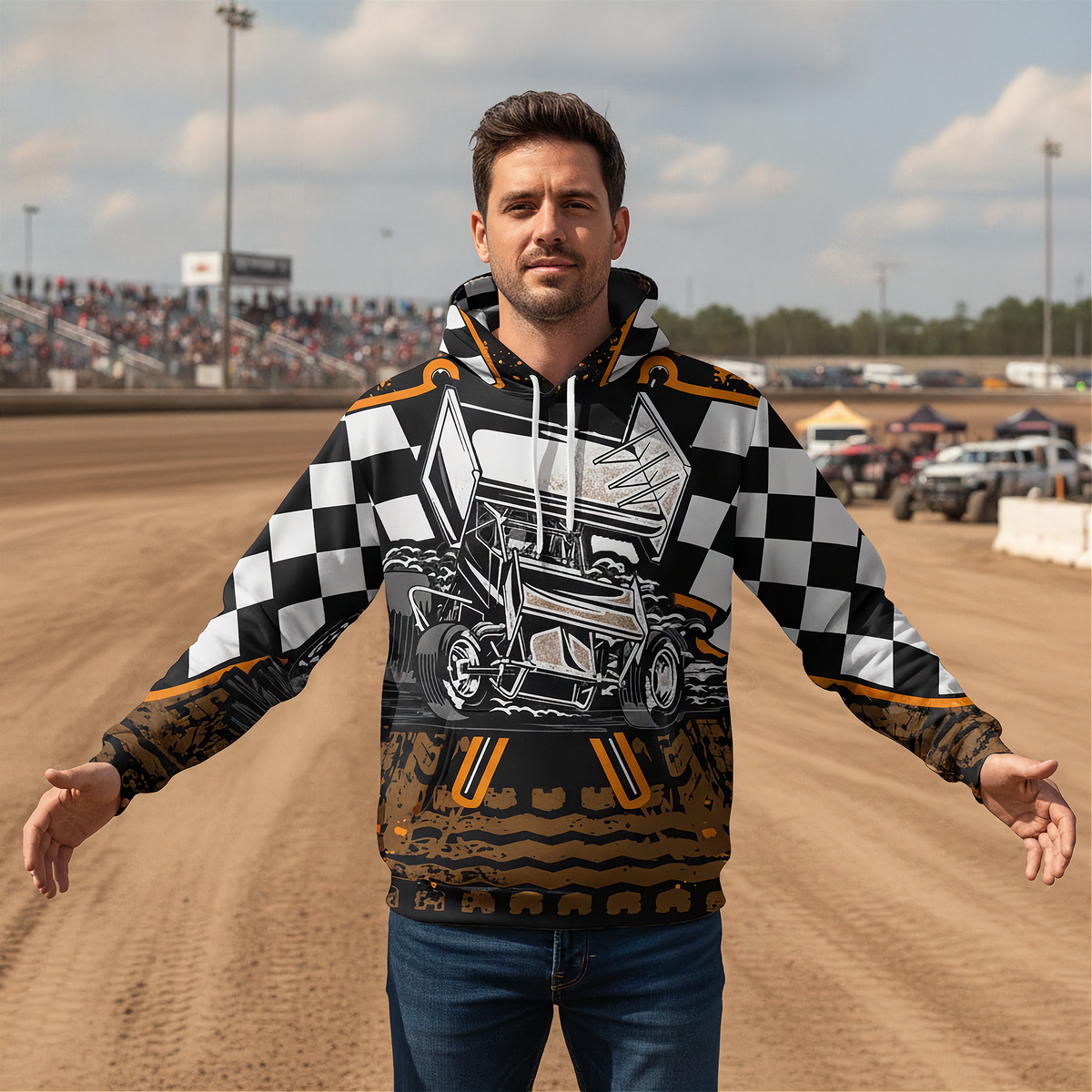 A man stands on a dirt race track wearing a graphic hoodie with a black-and-white checkered pattern, a white race car illustration on the chest, and brown and gold racing graphics near the hem. The background shows blurred race track bleachers, tents, and vehicles under a cloudy sky.