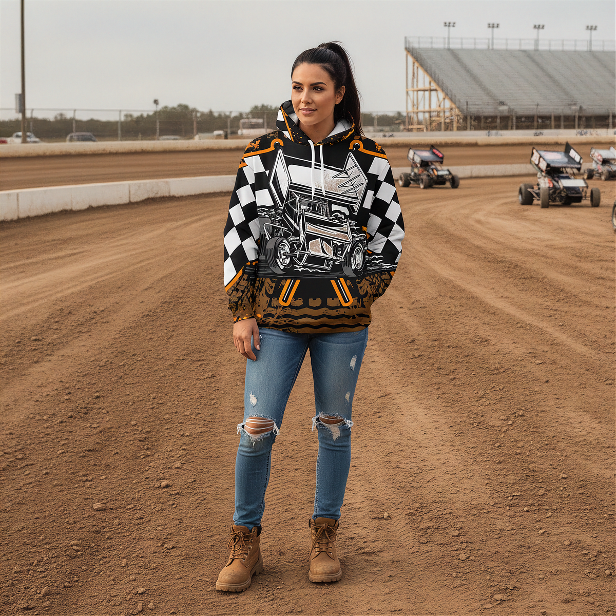 A woman stands on a dirt racetrack wearing a graphic hoodie with a black-and-white checkerboard pattern, a white race car illustration, and orange accents, paired with ripped blue jeans and brown boots. Racing vehicles and grandstands are visible in the background.