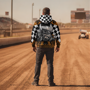 A man stands on a dirt racetrack facing away from the camera, wearing a black and white checkered hoodie with a large hot rod car graphic on the back, dark gray jeans, and black shoes. A race car is visible in the distance on the track.