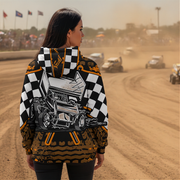 A woman stands on a dirt race track wearing a hooded jacket with a large back graphic of a race car, checkered shoulders, and racing-themed print. Dirt bikes and a blurred racetrack environment are visible in the background.