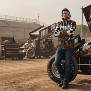 A man stands in a dirt track pit area wearing a black, white, and orange checkered hoodie with a tiger stripe pattern, blue jeans, and black shoes, with race cars and equipment visible in the background.