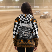 A person viewed from behind wearing a hooded sweatshirt with a race-track graphic and checkered pattern, standing on a dirt track with race cars blurred in the background.