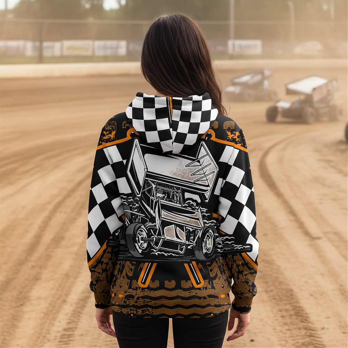 A person viewed from behind wearing a hooded sweatshirt with a race-track graphic and checkered pattern, standing on a dirt track with race cars blurred in the background.