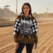 A woman stands on a dirt racetrack wearing a zip-up hoodie with a bold racing graphic. The hoodie features a black-and-white checkered pattern on the sleeves, a detailed race car illustration on the front, and orange and black graphic accents near the hem.