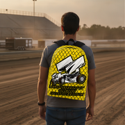 A man viewed from behind wearing a bright yellow backpack with a large white and black race car graphic and the words 'SPRINT CAR RACING' on the back, standing on a dirt racetrack at sunset.