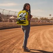 A woman stands on a dirt racetrack wearing a bright yellow and black patterned backpack with a large racing car graphic and the text 'Spring Car Racing'.