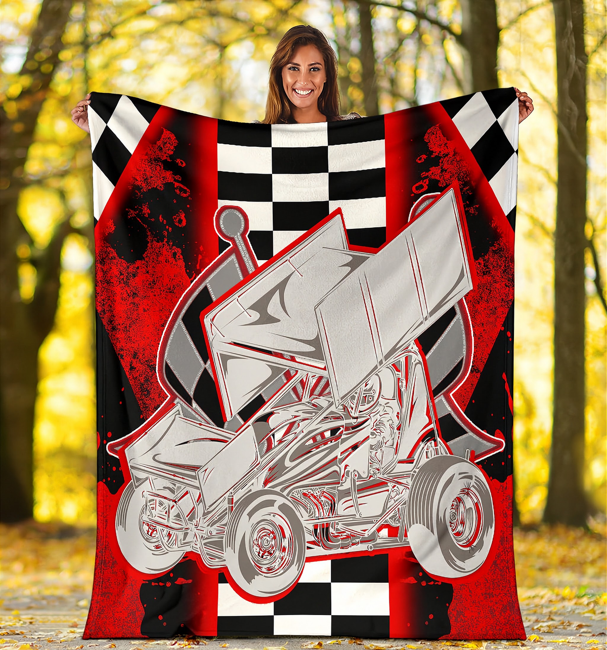 A woman stands outdoors in a wooded area holding up a large printed blanket or throw. The blanket features a bold graphic of a silver sprint car over a red, black, and white checkered background with distressed splatter effects.