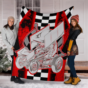 Two women holding up a large fleece blanket featuring a graphic of a silver sprint car on a red and black background with a checkered flag pattern. The setting is outdoors in front of a white house door with snow on the ground.