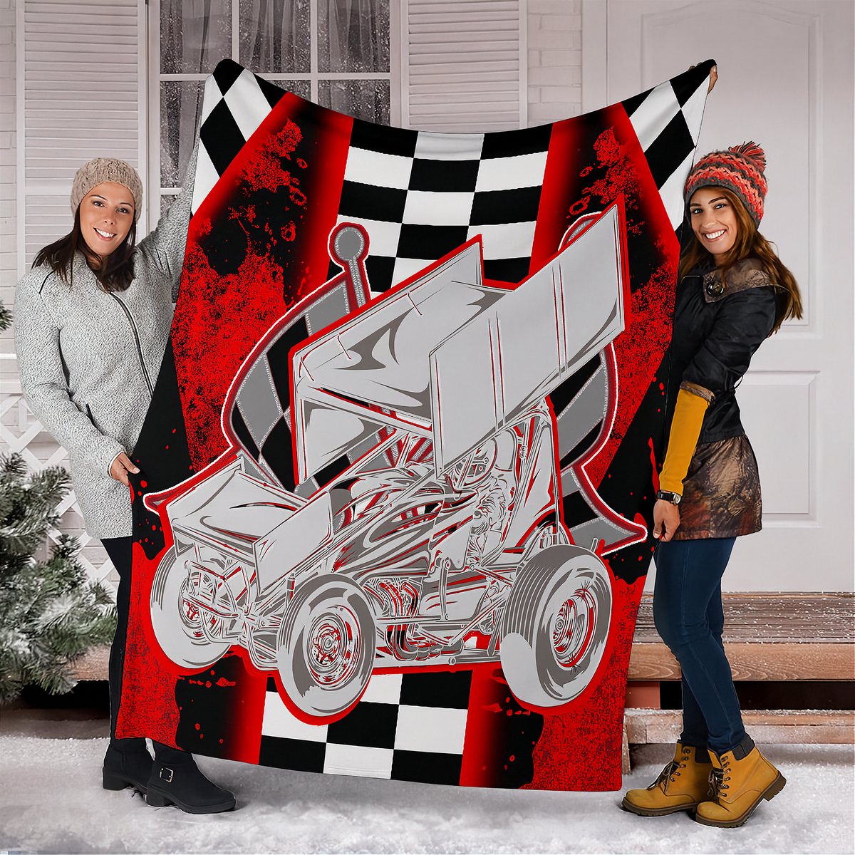 Two women holding up a large fleece blanket featuring a graphic of a silver sprint car on a red and black background with a checkered flag pattern. The setting is outdoors in front of a white house door with snow on the ground.