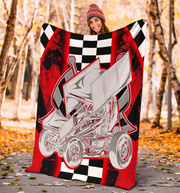 A person wearing a knit beanie holds up a large blanket or throw in an outdoor autumn setting with trees and fallen leaves. The blanket features a bold graphic design with a silver sprint car over a red, black, and white checkered background.