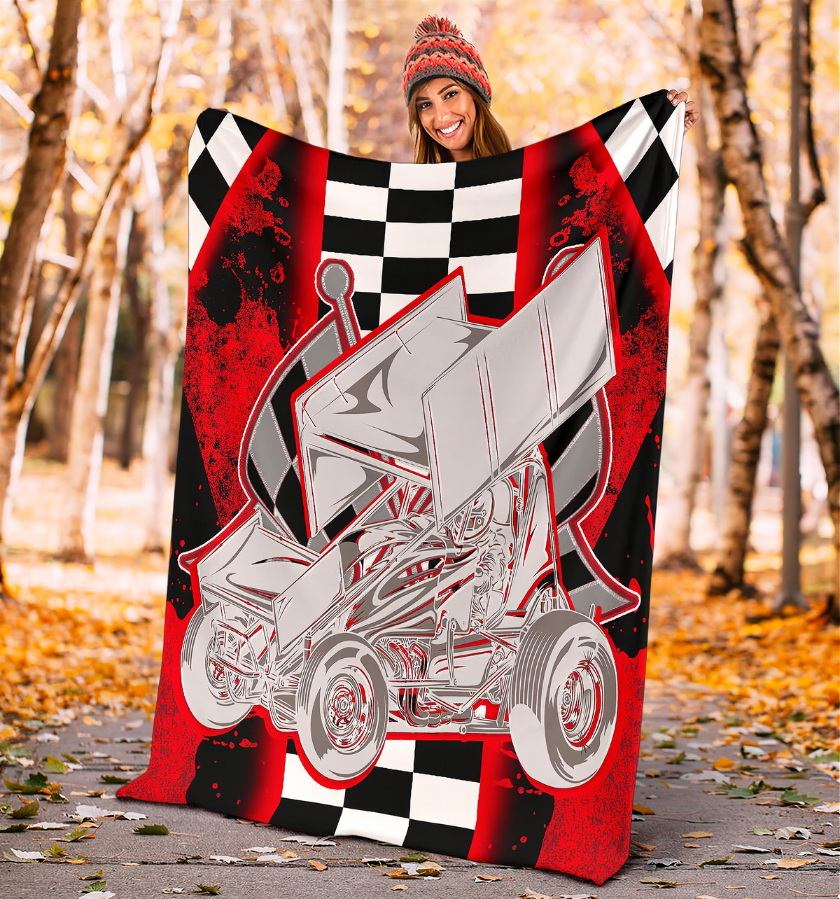 A person wearing a knit beanie holds up a large blanket or throw in an outdoor autumn setting with trees and fallen leaves. The blanket features a bold graphic design with a silver sprint car over a red, black, and white checkered background.