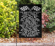 A black rectangular garden flag with white racing-themed prayer text and crossed checkered flags at the top, displayed on a black metal stand in a garden with green shrubs and mulch.