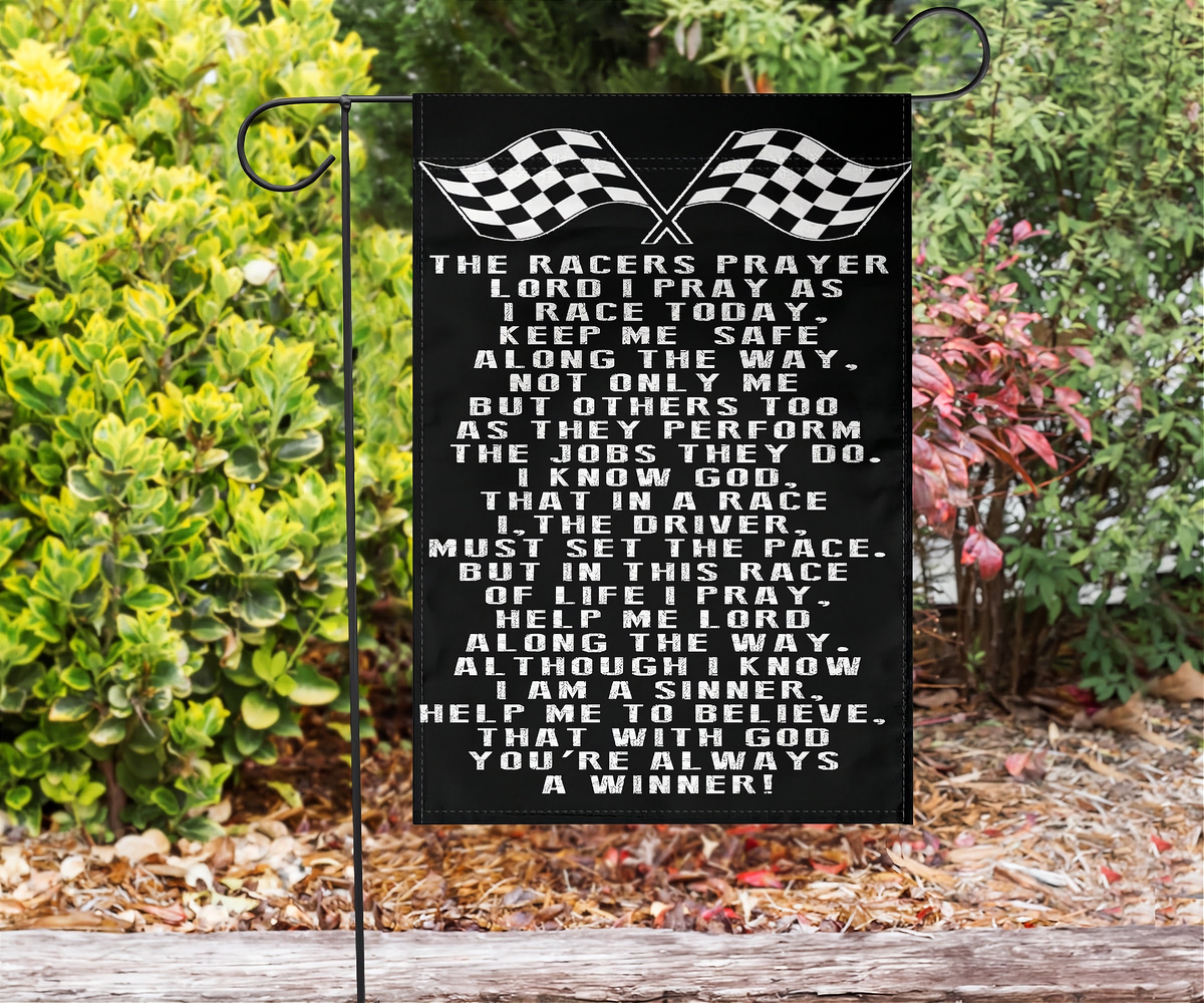 A black rectangular garden flag with white racing-themed prayer text and crossed checkered flags at the top, displayed on a black metal stand in a garden with green shrubs and mulch.