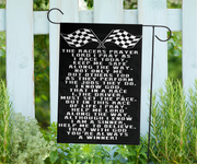 A black garden flag with white text and two checkered flags at the top, hanging from a black metal stand outdoors in front of a white fence and green plants.