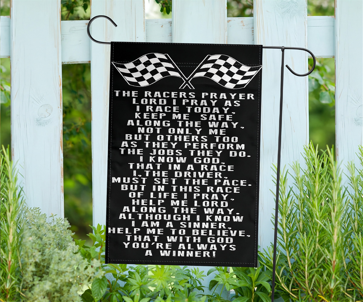 A black garden flag with white text and two checkered flags at the top, hanging from a black metal stand outdoors in front of a white fence and green plants.