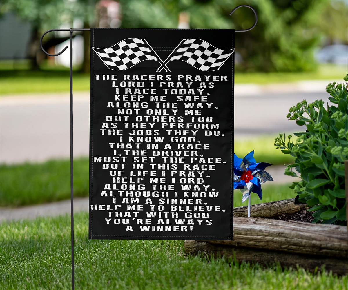 A black rectangular garden flag with white racing-themed text and two checkered flags at the top, displayed on a metal stand in a grassy outdoor setting next to a small blue and red wind spinner.