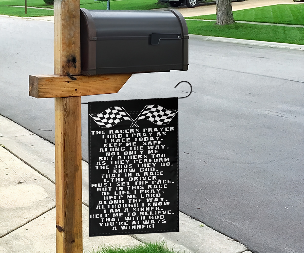A black metal hanging sign with white racing-themed text and checkered flags, mounted on a wooden post next to a mailbox on a suburban sidewalk.