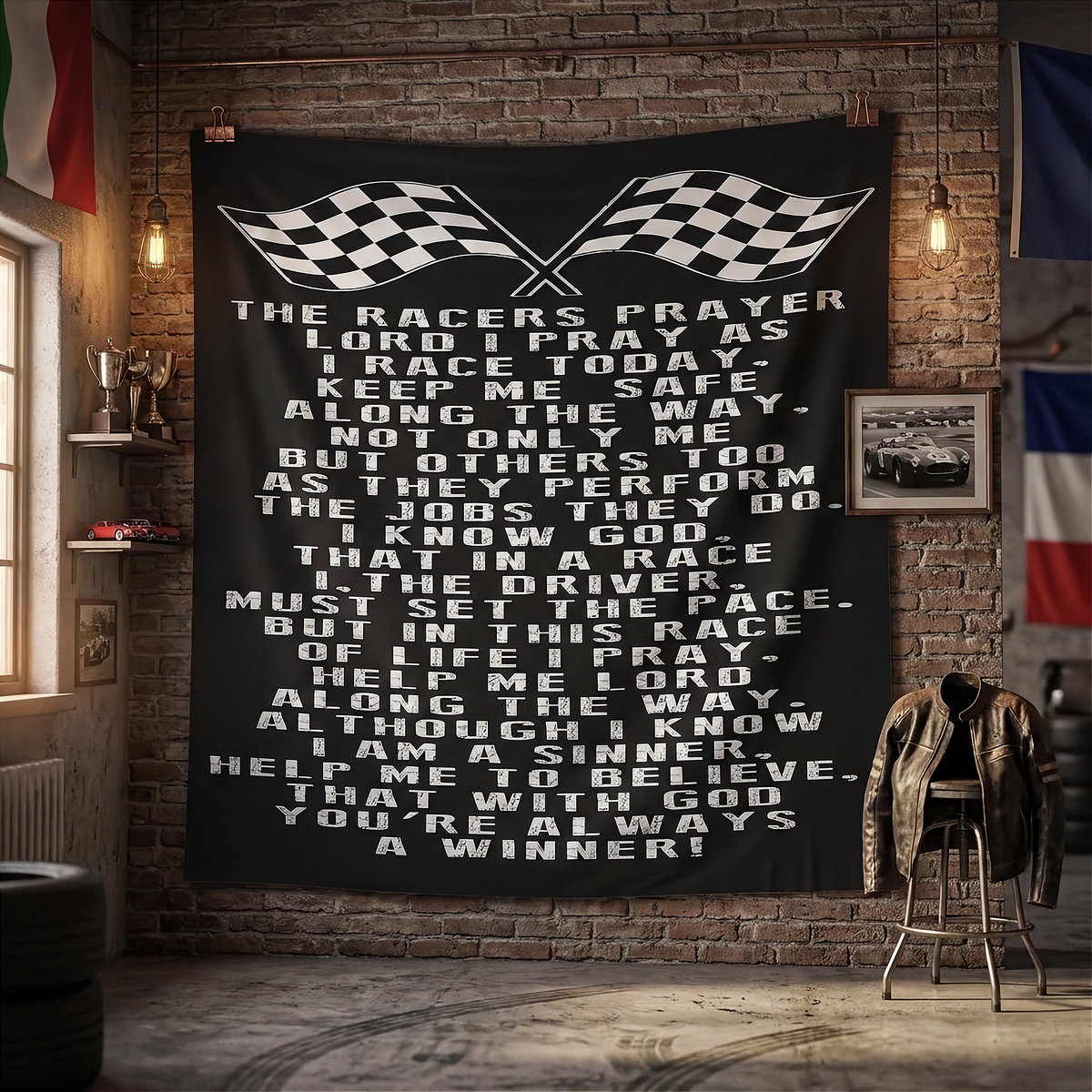 Black Racer’s Prayer tapestry with white lettering and crossed checkered flags hanging on a brick wall beside trophies and a racing jacket.