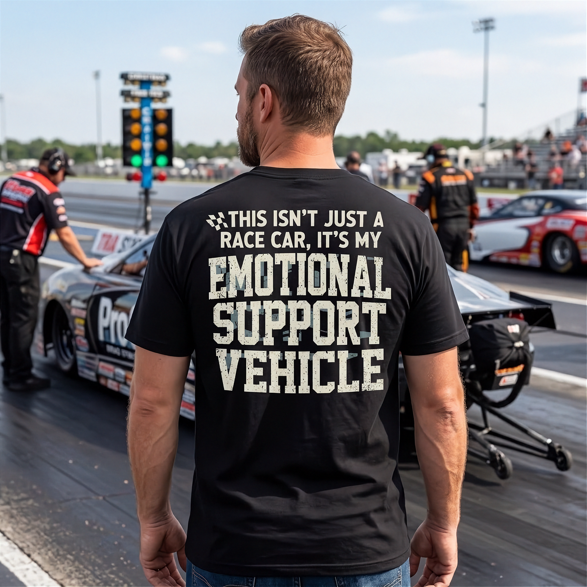 A man standing on a racetrack wearing a black t-shirt with a humorous graphic printed on the back. The text reads: 'THIS ISN'T JUST A RACE CAR, IT'S MY EMOTIONAL SUPPORT VEHICLE' in distressed white lettering.
