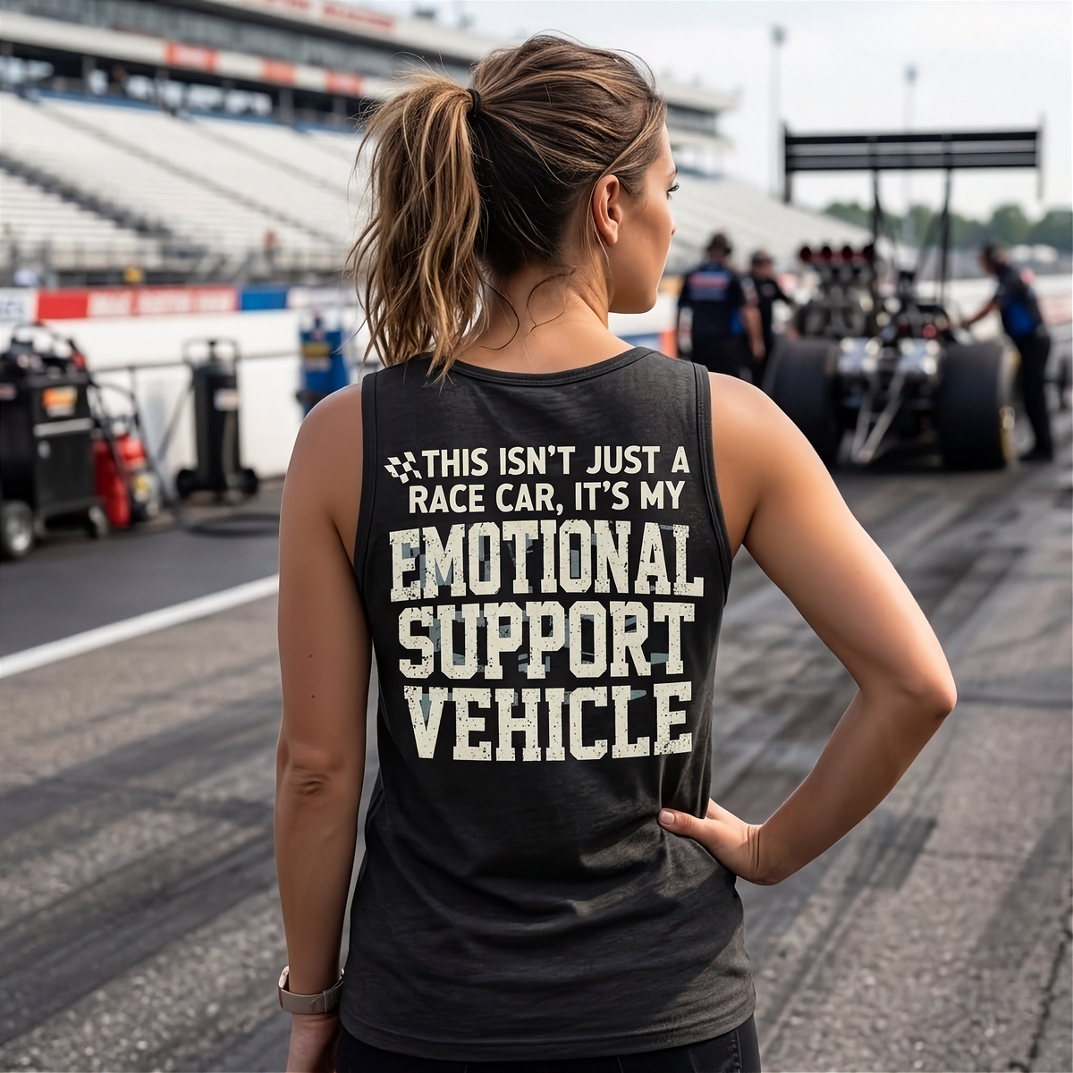 A woman stands on a racetrack wearing a dark gray racerace tank top with the text 'THIS ISN'T JUST A RACE CAR, IT'S MY EMOTIONAL SUPPORT VEHICLE' printed in distressed white letters on the back.