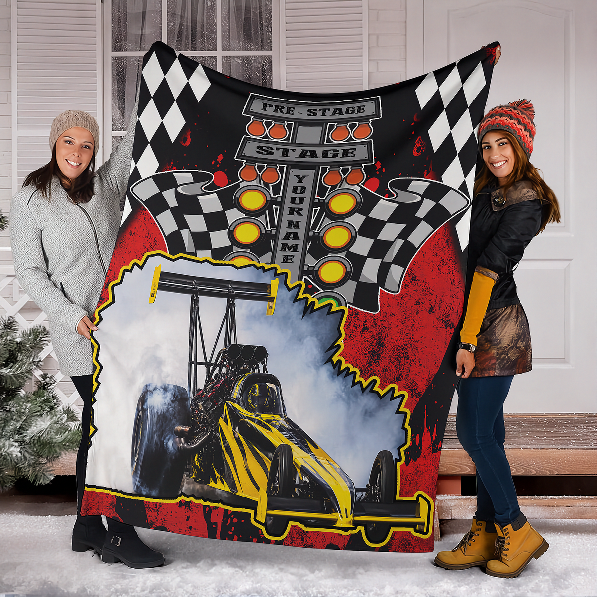 Two women holding a personalized dragster blanket outside a snowy home, featuring a yellow-and-black car and sample YOUR NAME text.