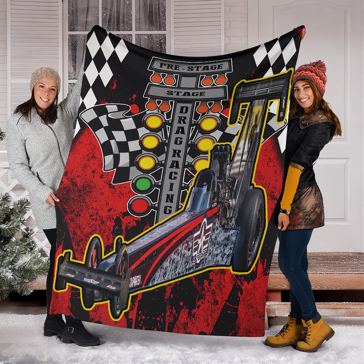Two women holding a red-and-black dragster blanket outside a snowy home, showing the full car photo and starting-light design.