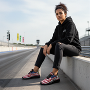 Woman sitting on a race track wearing black hoodie and pants with usa flag drag racing shoes.
