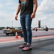 man standing on a race track wearing a black t-shirt with a graphic design, blue jeans, and American flag drag racing sneakers. In the background, there is a classic car and traffic lights.