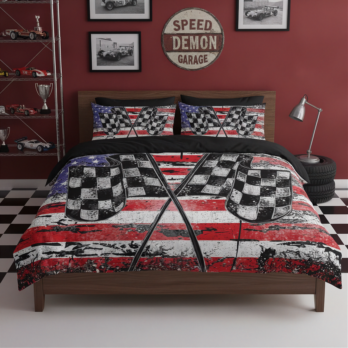 A bed dressed in a racing-themed bedding set with a distressed American flag pattern, checkered flags, and black-and-white checkered pillows, shown in a styled garage room with red walls, framed pictures, a shelf with toy cars, and a lamp.