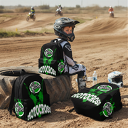 A child wearing motocross gear sits on a tire at a dirt track, with three black and green motocross backpacks around them. One backpack is worn on the child's back, and two are placed on the ground.