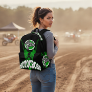 A woman stands on a dirt track wearing a black backpack with green and white motocross graphics and the word 'MOTOCROSS' printed across the back.