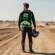 A man standing on a dirt motocross track, viewed from behind, wearing a black and green motocross jersey with 'MOTOCROSS' printed across the back, black pants, knee pads, and holding a helmet.