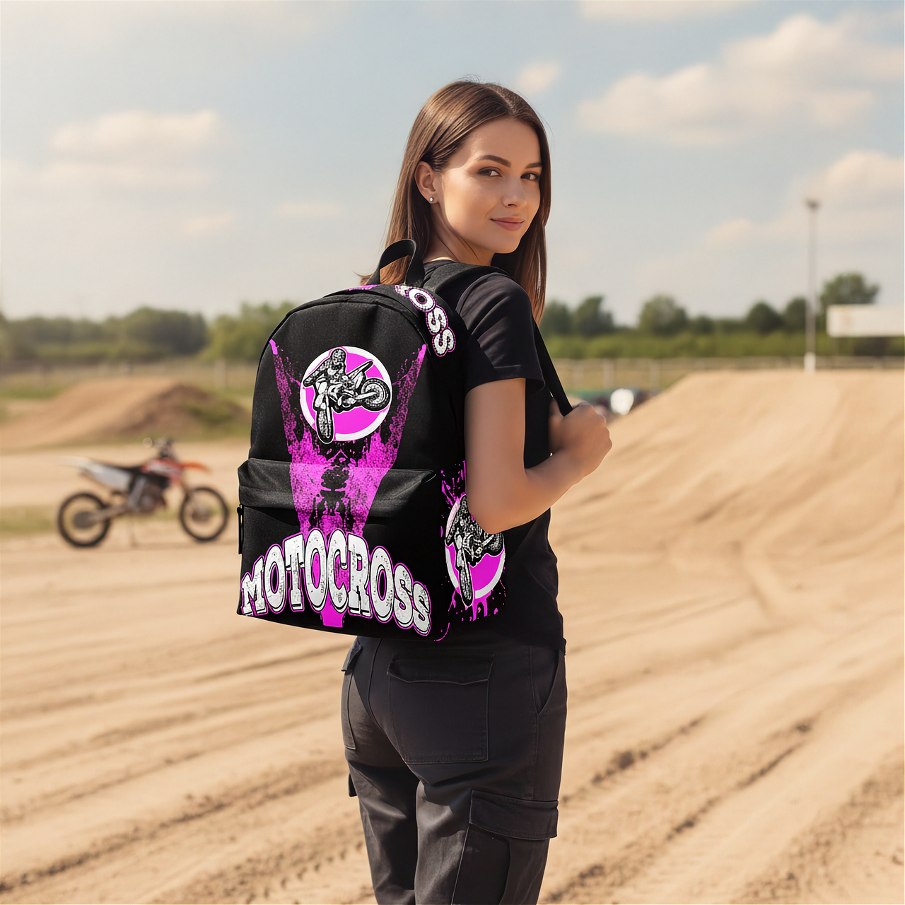 A woman stands on a dirt track wearing a black backpack with a large pink and white motocross graphic on the back. A motocross bike is visible in the background under a cloudy sky.