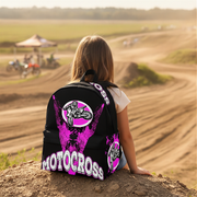 A person seen from behind sitting on a dirt mound at a motocross track, wearing a black backpack with bright pink and white graphics that read 'MOTOCROSS' and feature a motocross rider logo.