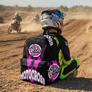 A motocross rider sits on a dirt track wearing a black, neon green, and pink printed jersey and helmet, with two riders kicking up dust in the background.