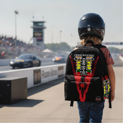 A child wearing a black racing helmet and a black backpack with a large red and yellow 'Prestige Racing' graphic stands on a racetrack, viewed from behind.