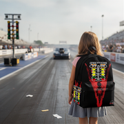 A girl stands on a racetrack facing a race car, wearing a black backpack with a large red, yellow, and white racing graphic on the back.