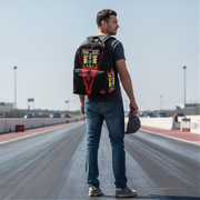 A man standing on a racetrack wearing a black backpack with a large red, yellow, and white graphic design on the back, a dark gray t-shirt, and blue jeans.