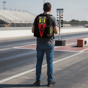 A man standing on a racetrack seen from behind, wearing a black drag racing backpack with a large red, yellow, and white graphic design on the back.