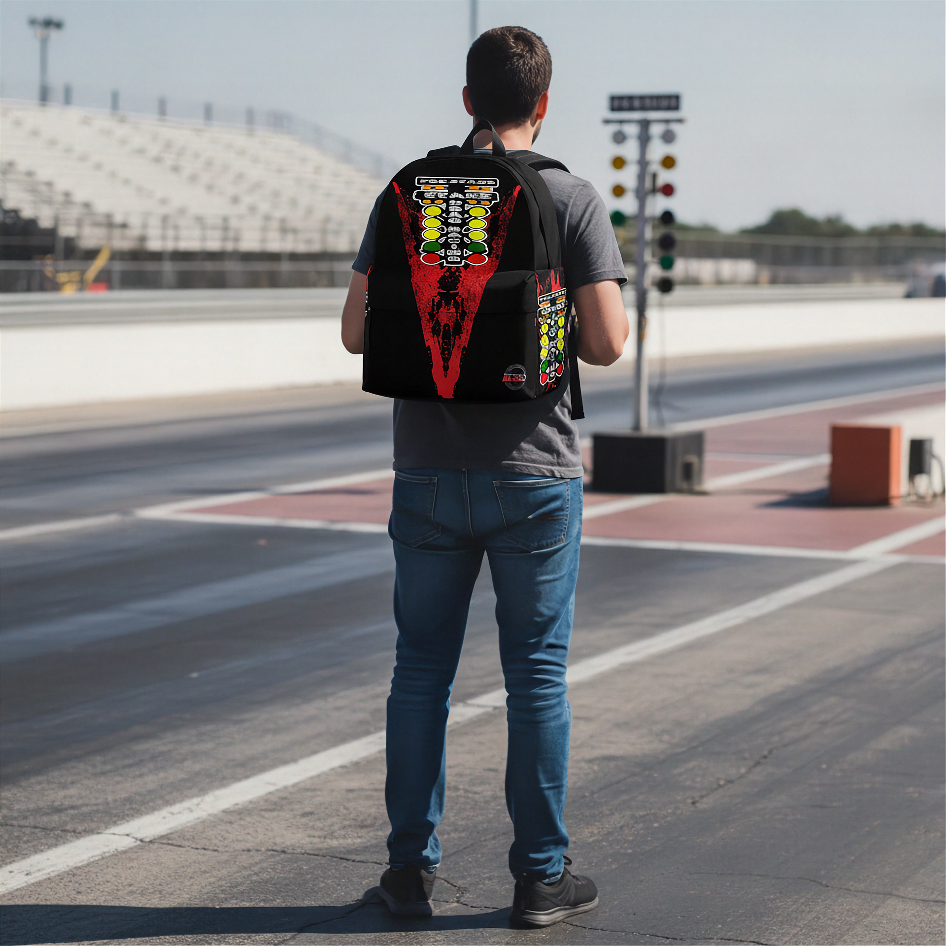 A man standing on a racetrack seen from behind, wearing a black drag racing backpack with a large red, yellow, and white graphic design on the back.
